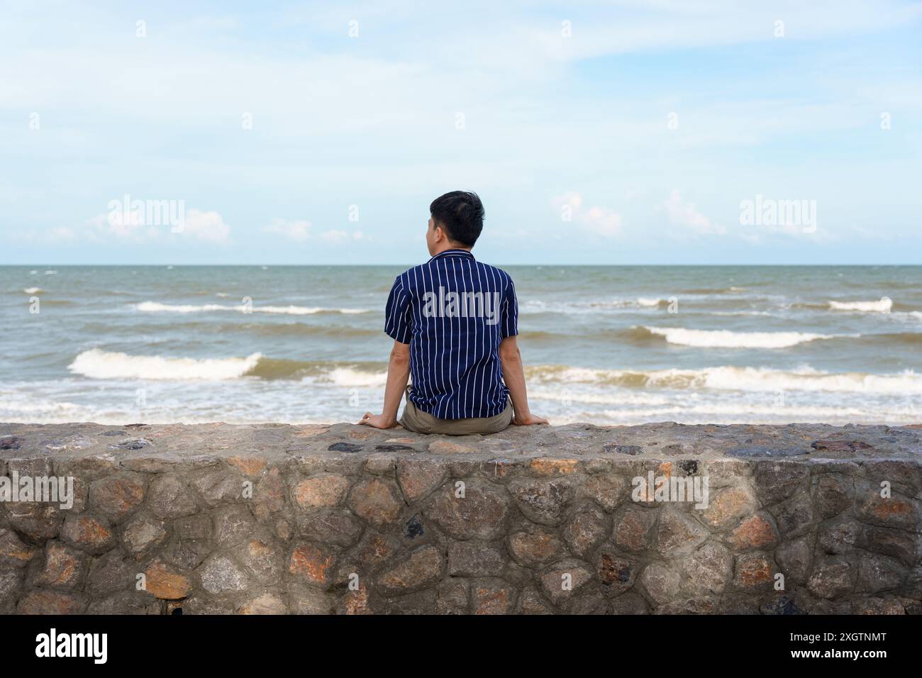 Rückansicht eines jungen asiatischen Mannes, der am Sommerwochenende auf der Promenade im tropischen Meer sitzt Stockfoto