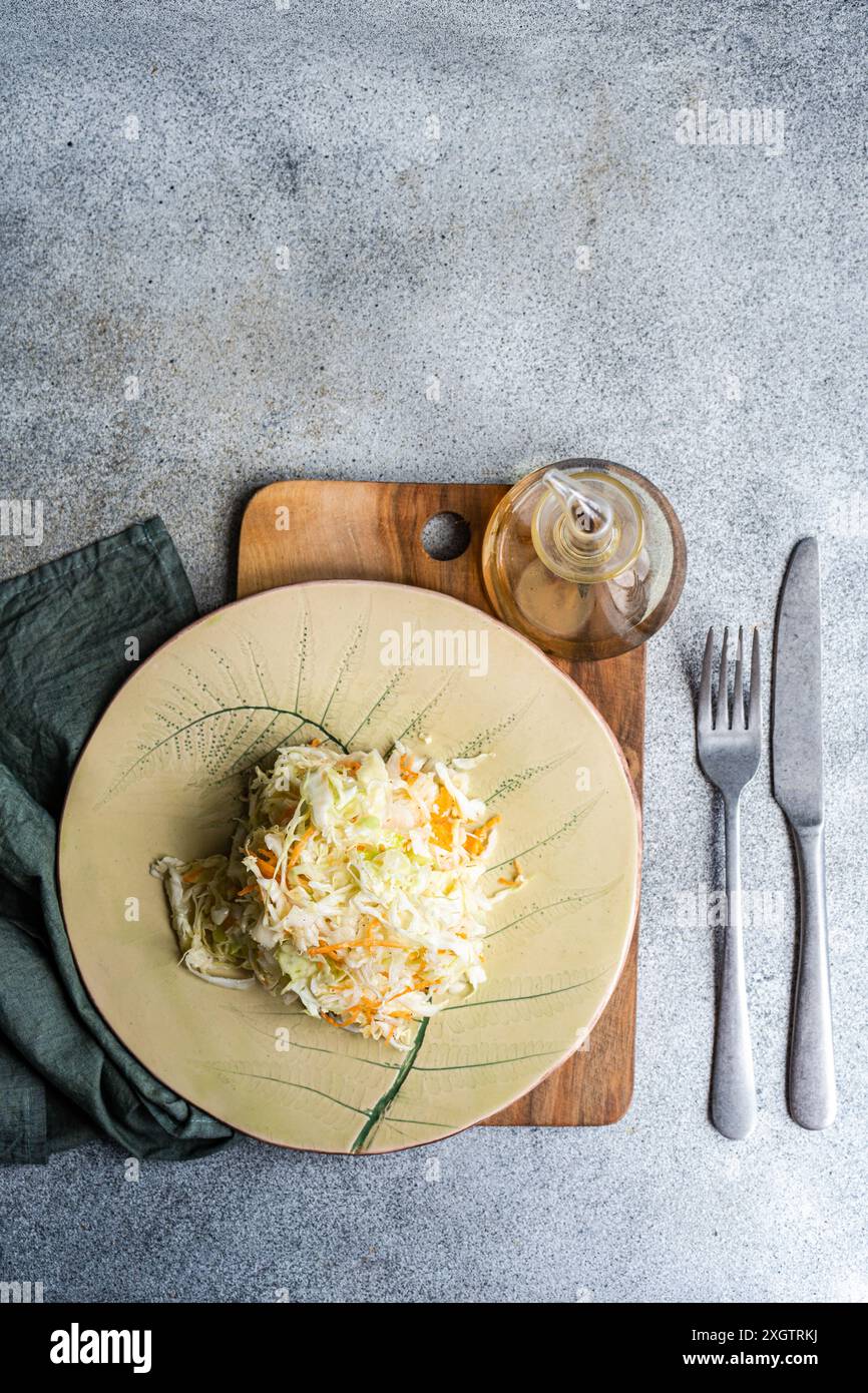 Blick von oben auf einen nahrhaften Bio-Kohl- und Karottensalat mit Olivenöl, präsentiert auf einer Keramikplatte, begleitet von einer Gabel, einem Messer und einem g Stockfoto