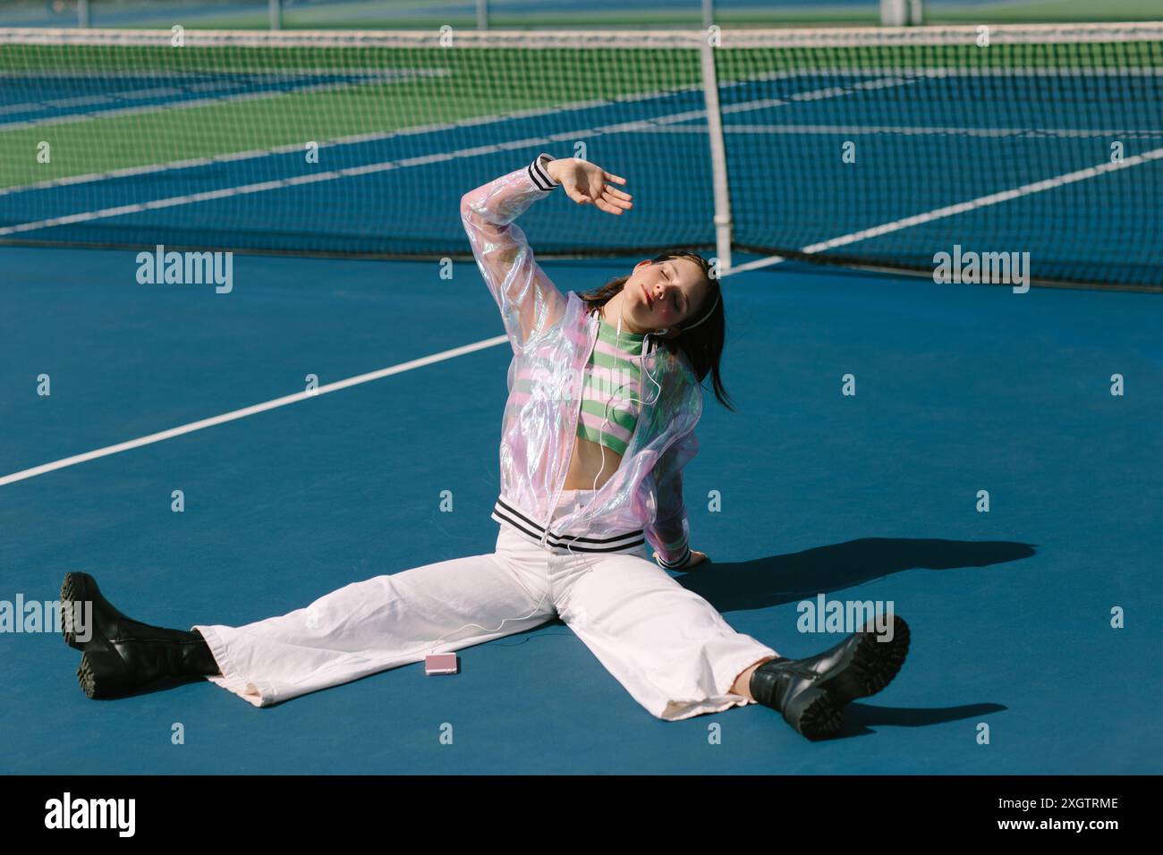 Eine junge Frau, die eine reflektierende transparente Jacke und weiße Hose trägt, ist in einem fröhlichen Moment gefangen, während sie Musik auf einem blauen Tennisplatz H hört Stockfoto