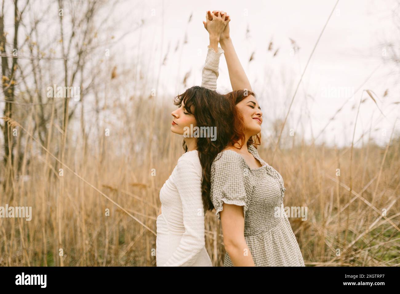 Zwei junge Frauen in Vintage-Kleidern stehen Rücken an Rücken, Hände geschlossen und Augen geschlossen, und genießen einen ruhigen Moment in einer natürlichen, bewaldeten Umgebung Stockfoto