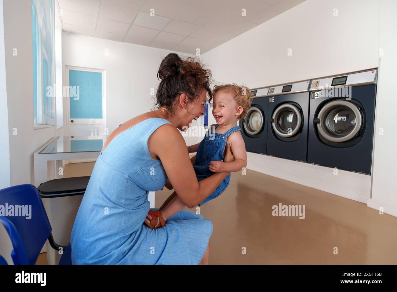 Hispanische Frau spielt mit ihrem Sohn, während sie wartet, bis die Wäsche im Waschsalon fertig ist. Selbstbedienungs-Waschsalon. Stockfoto