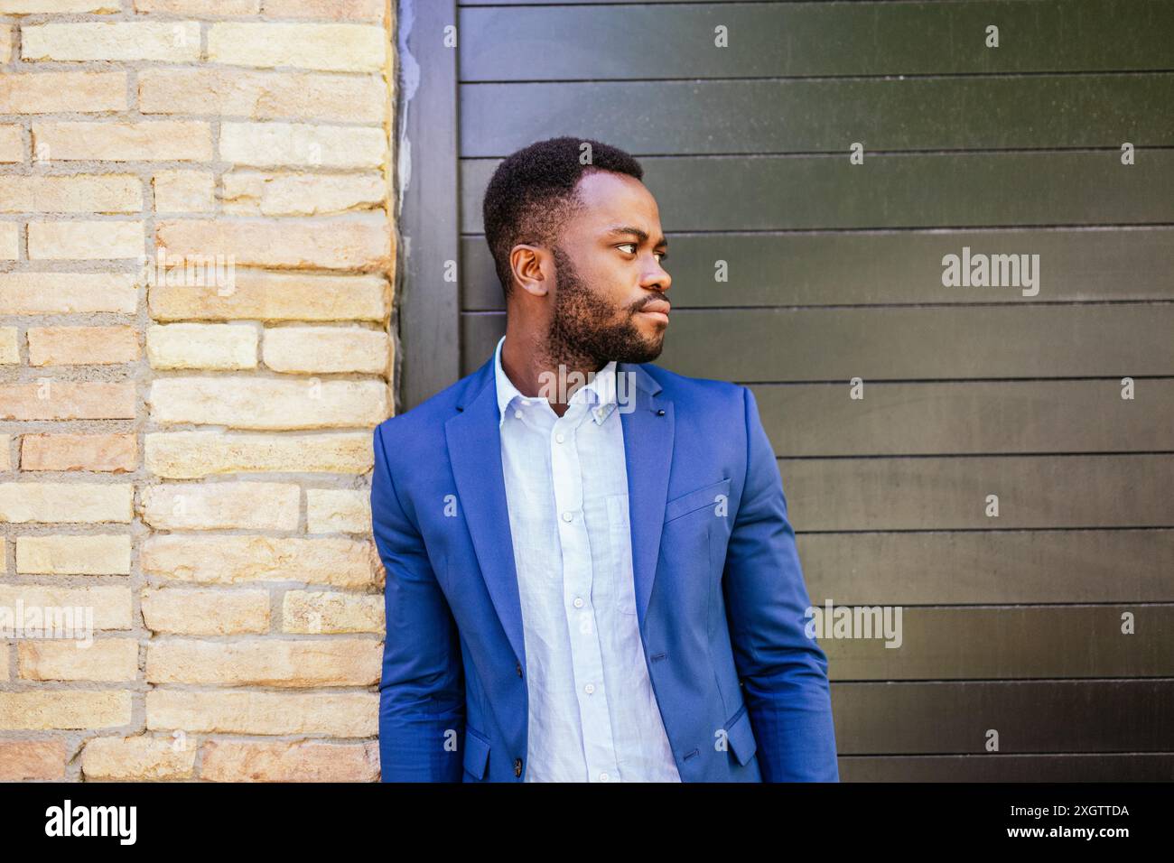 Ein Porträt eines besinnlichen jungen afroamerikanischen Mannes in einem stilvollen blauen Anzug, der vor einem strukturierten Backstein- und Metallhintergrund blickt. Stockfoto
