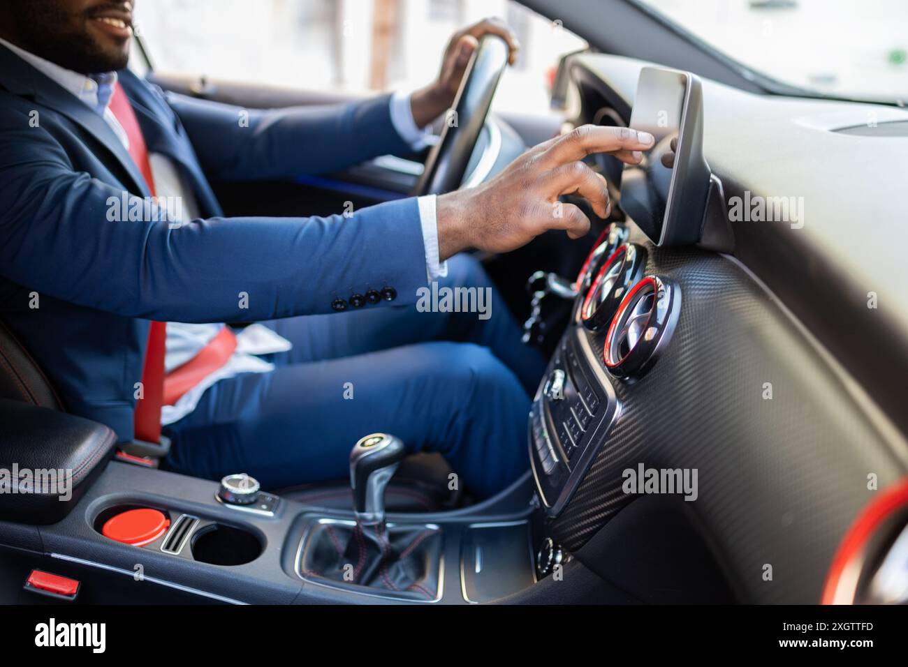 Ein afroamerikanischer Geschäftsmann, der in einem blauen Blazer gekleidet ist, passt den Touchscreen eines modernen Autos an, während er lächelt. Stockfoto