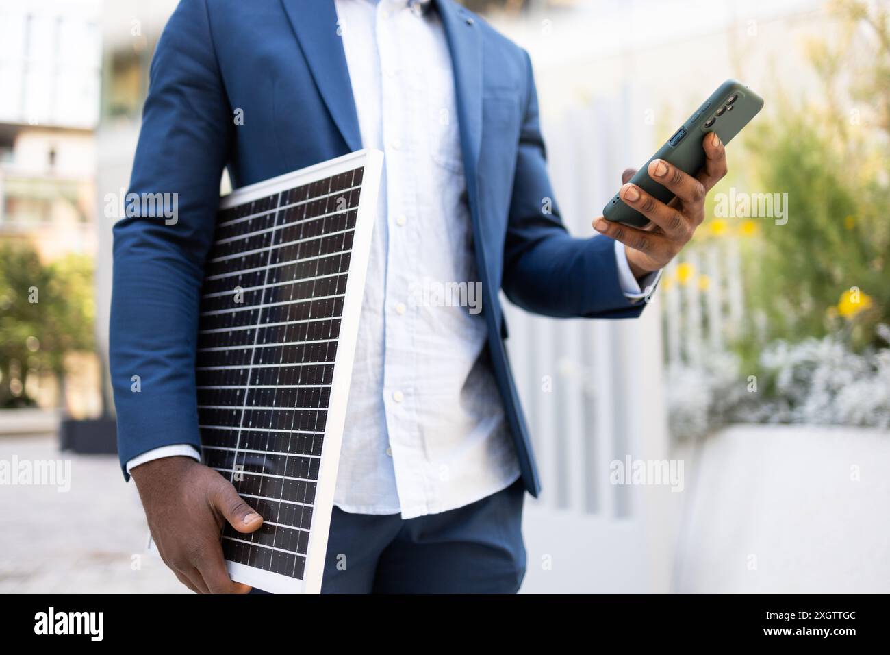 Schwarzer Geschäftsmann, der stilvoll gekleidet ist und ein Solarpanel und ein Smartphone hält und sich intensiv auf sein Gerät konzentriert. Stockfoto