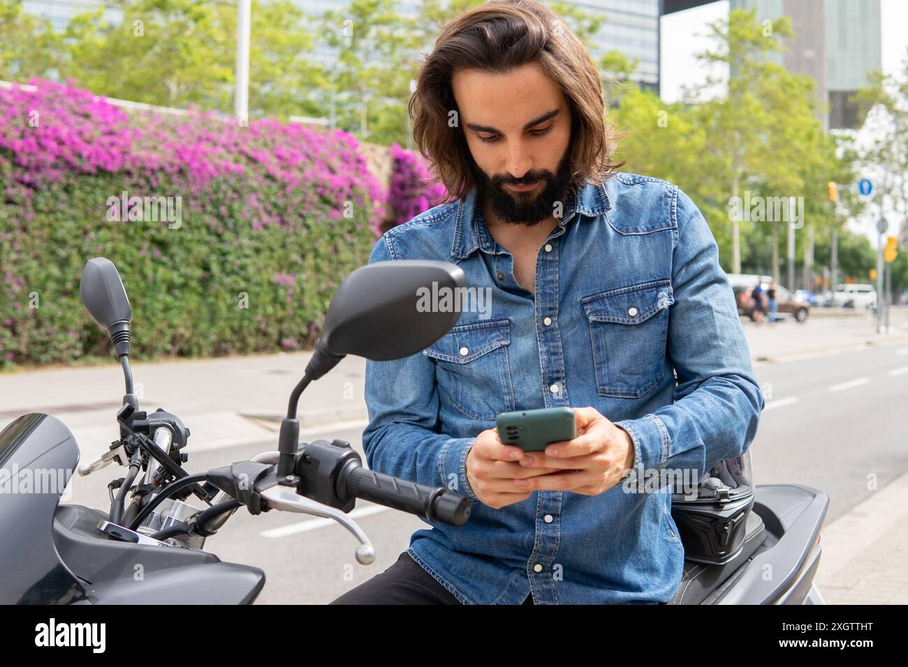 Ein junger Mann mit langen Haaren und einer jeansjacke überprüft sein Smartphone, während er auf einem Roller im lebhaften Stadtteil Poblenou sitzt und die Moderne präsentiert Stockfoto