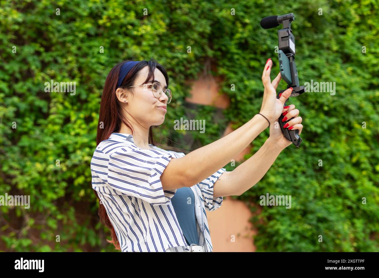 Eine fröhliche junge Frau mit einem mobilen Gimbal vloggt in einem üppigen Garten und nimmt Inhalte mit einem Smartphone auf Stockfoto