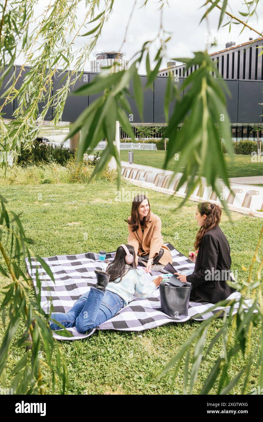Drei Studenten sitzen auf einer karierten Decke in einem üppigen Stadtpark, vertiefen sich in Gesprächen und genießen die Gesellschaft des anderen, umgeben von urbaner Architektur Stockfoto
