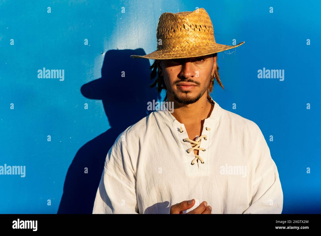 Ein junger Kaukasier mit Dreadlocks und Strohhut steht vor einem leuchtend blauen Hintergrund. Er blickt direkt in die Kamera mit einem selbstbewussten Ausdruck Stockfoto