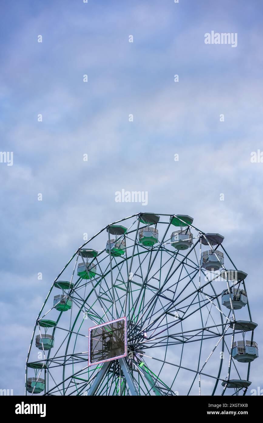 Ein Bild, das ein großes Riesenrad mit grünen und weißen Kabinen vor einem teilweise bewölkten blauen Himmel zeigt. Die Lichter des Gebäudes verleihen ein dezentes Leuchten. Stockfoto