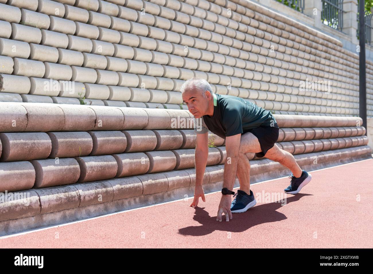 Ein älterer männlicher Athlet hockt sich in der Startposition auf einer roten Laufbahn, bereit zum Sprinten, was Gesundheit und Vitalität im späteren Leben Agave veranschaulicht Stockfoto