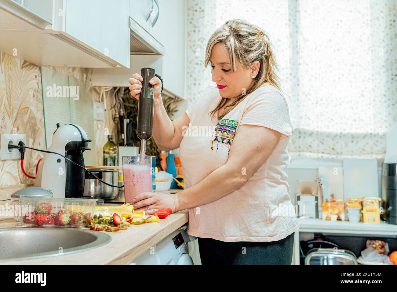 Eine Frau konzentriert sich auf das Mischen eines rosa Smoothies mit einem Stabmixer in ihrer überdachten Küche, umgeben von frischem Obst und Küchengeräten Stockfoto