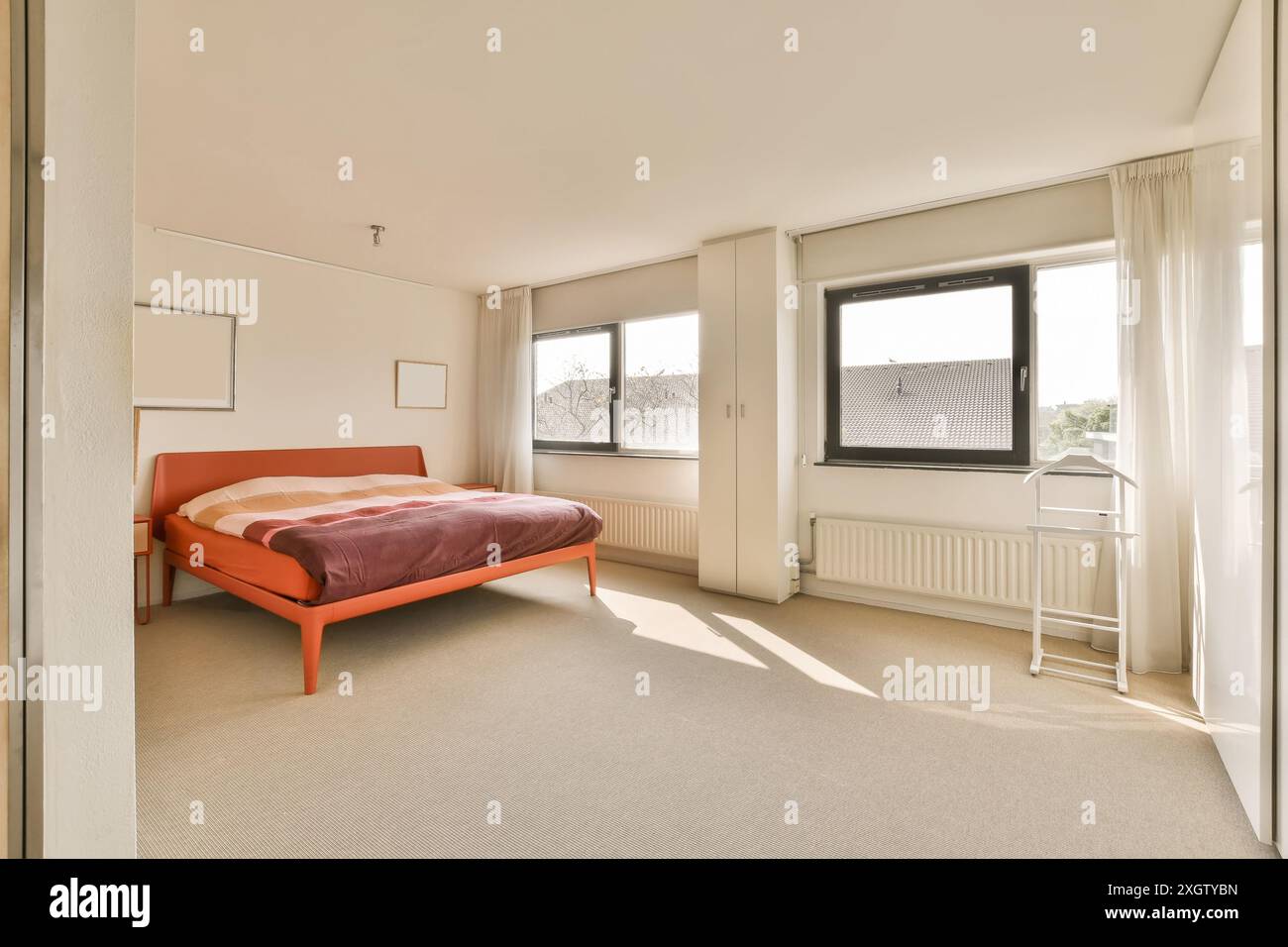 Modernes und luftiges Schlafzimmer mit einem großen Fenster mit Sonnenlicht in der Herentalsstraat, Amsterdam. Das Zimmer verfügt über ein orangefarbenes Bett, weiße Vorhänge und eine Meile Stockfoto