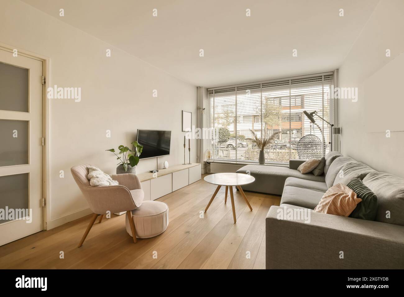 Geräumiges, modernes Wohnzimmer mit komfortablem Schlafsofa, minimalistischem TV-Ständer, stilvollem Sessel mit Polsterhocker und großem Fenster für Vormittag Stockfoto