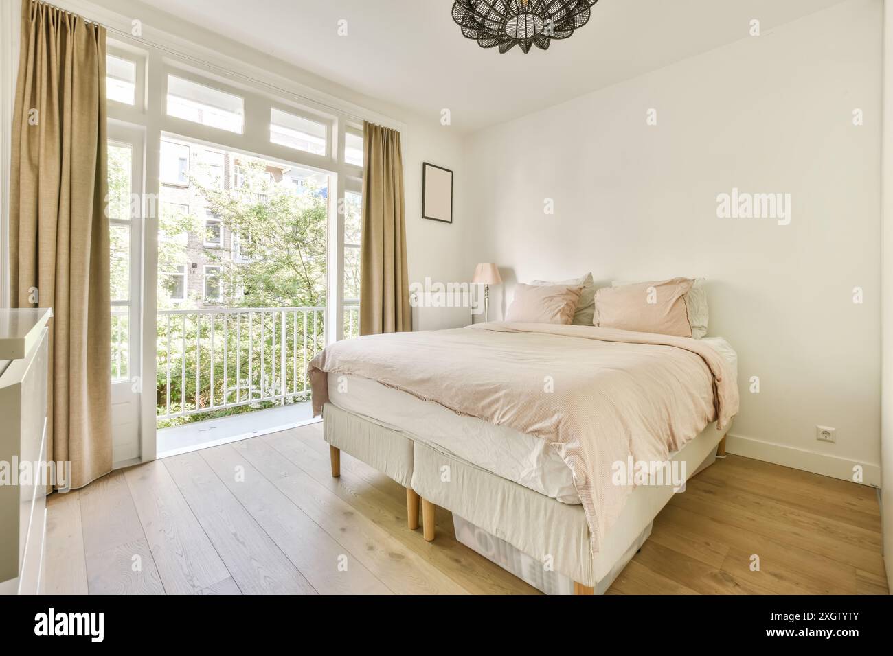 Ein gut beleuchtetes Schlafzimmer mit großem Fenster, Holzfußboden und Zugang zu einem Balkon, minimalistisches Design und helle Farbtöne. Stockfoto