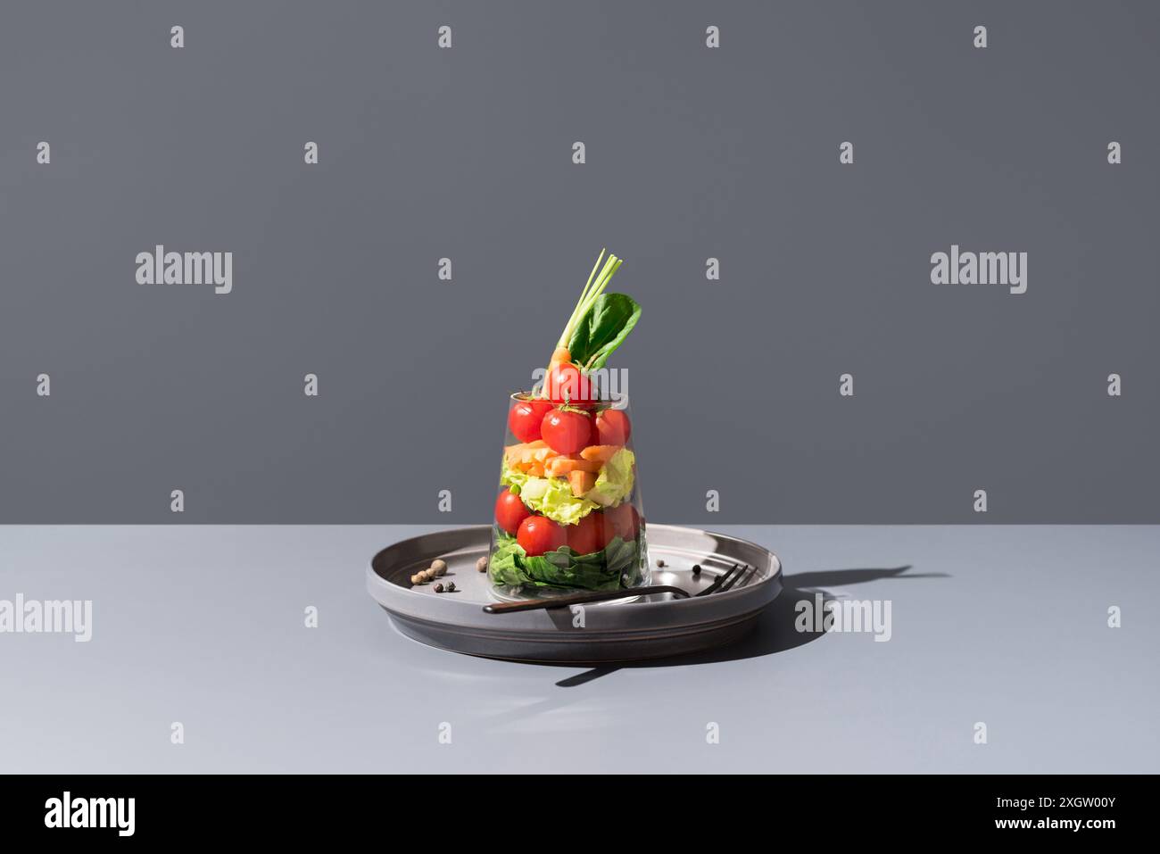 Kreativ gestufter Salat mit Tomaten, Karotten und Salat in einem transparenten Glas, elegant auf einem grauen Hintergrund mit scharfen Schatten. Stockfoto