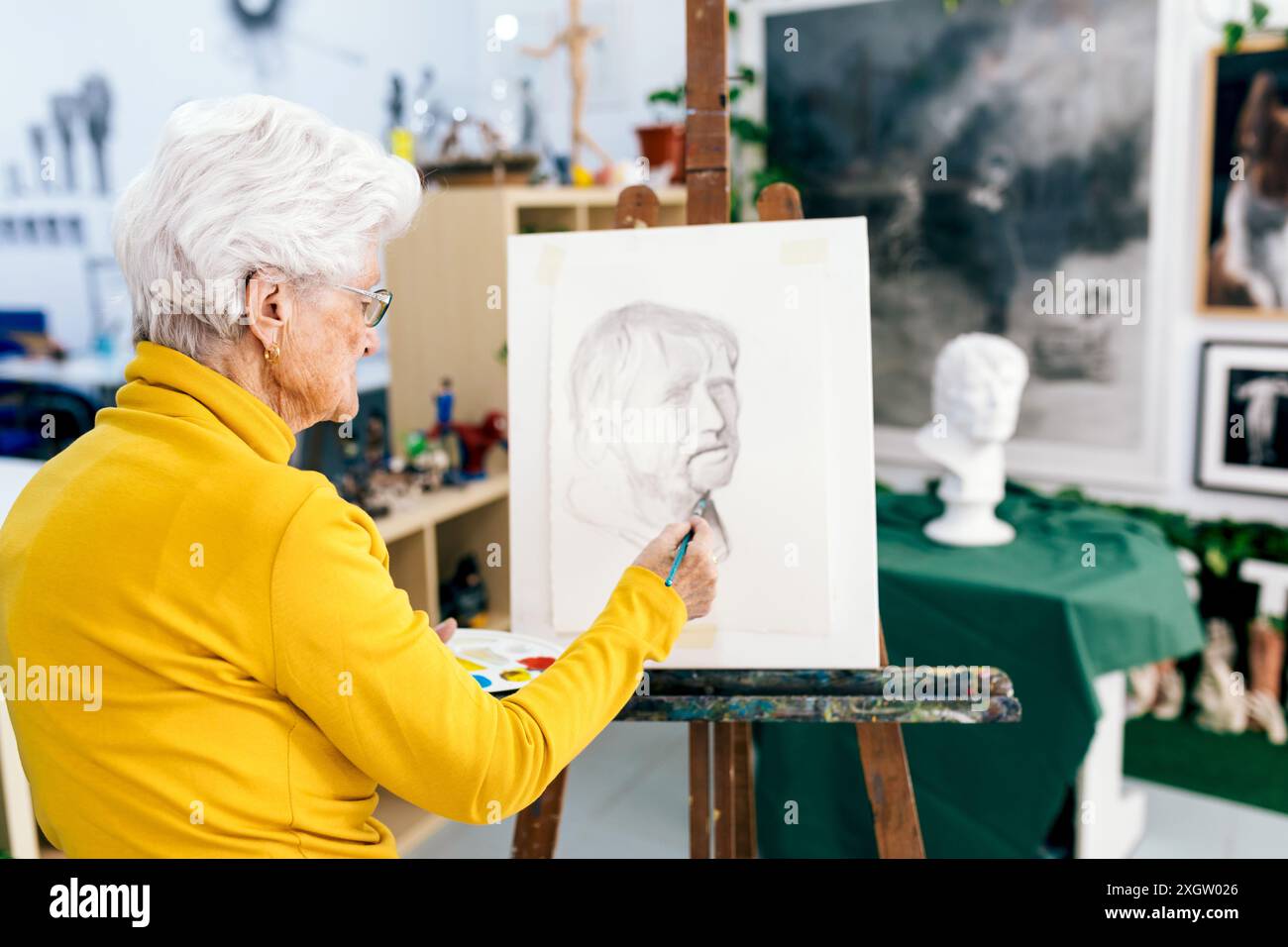 Eine ältere Frau mit weißen Haaren, die in einen gelben Pullover gekleidet ist, konzentriert sich auf das Malen eines Porträts auf Leinwand in einem gut beleuchteten Kunststudio, umgeben von verschiedenen Stockfoto