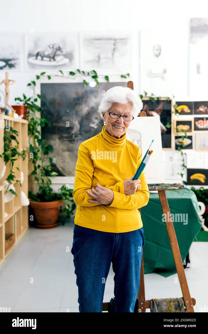 Eine fröhliche ältere Künstlerin steht in ihrem gut beleuchteten Kunststudio mit Pinseln, umgeben von üppigen Pflanzen und Kunstwerken Stockfoto
