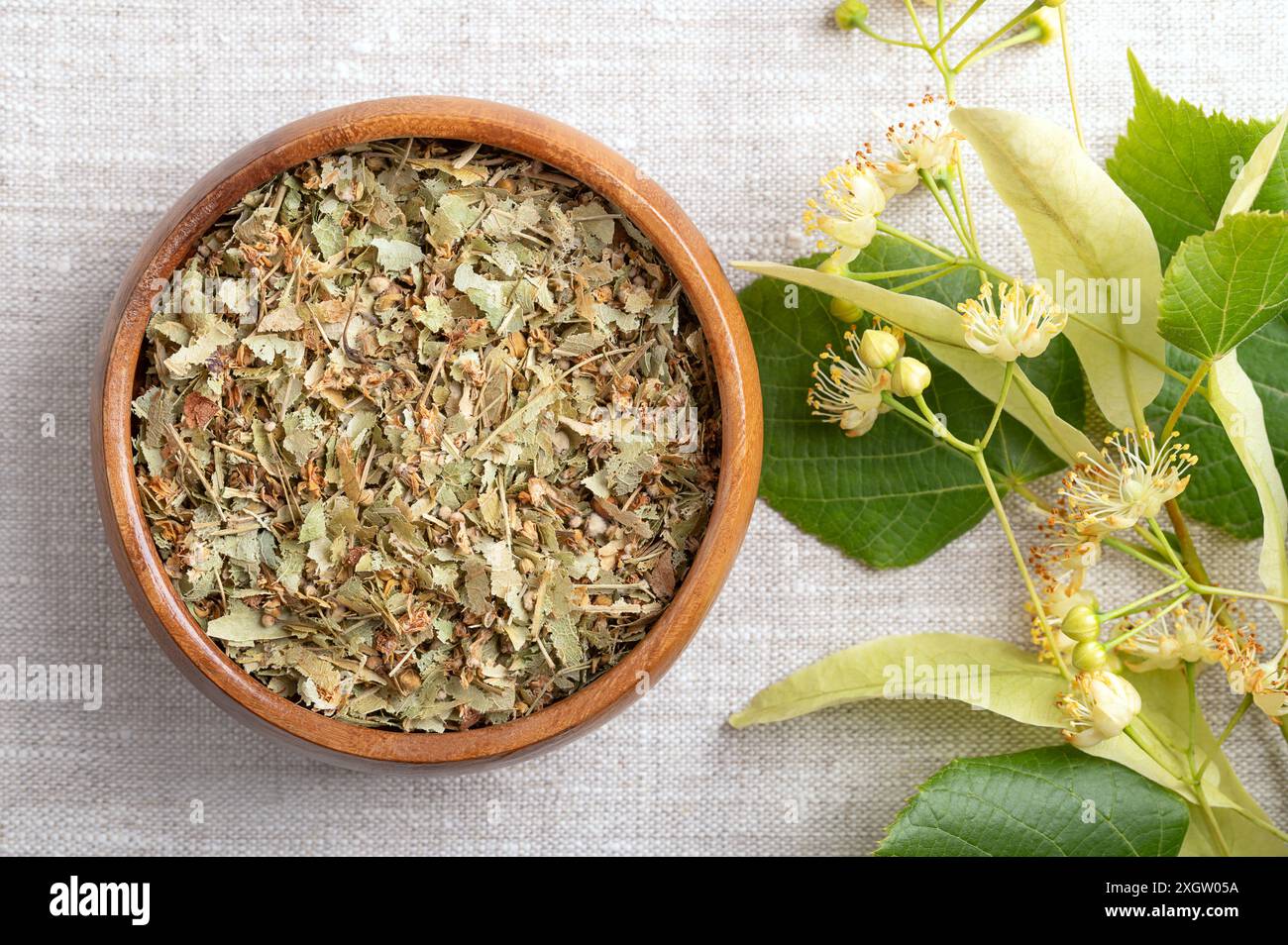 Linden blüht in einer Holzschale auf Leinenstoff. Getrocknete, frische Blüten, Früchte und Blätter der großblättrigen Linde oder Linde, Tilia platyphyllos. Stockfoto
