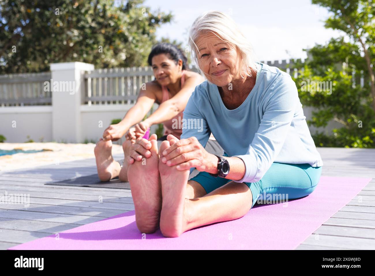 Zwei glückliche ältere Freundinnen, die Yoga machen, die Beine ausstrecken und im sonnigen Garten sitzen. Yoga, Wohlbefinden, Bewegung, Freundschaft und gesunde Senioren Stockfoto