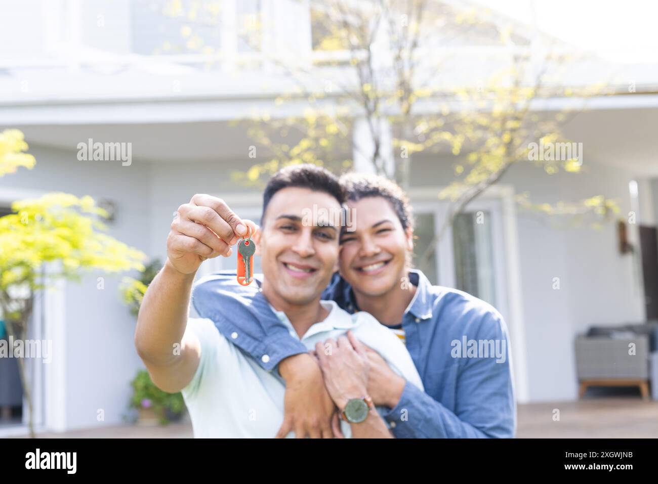 Ein vielseitiges Paar zeigt stolz neue Hausschlüssel. Sie feiern unveränderte Eigenheimbesitzer im Freien in einer sonnigen Wohngegend. Stockfoto