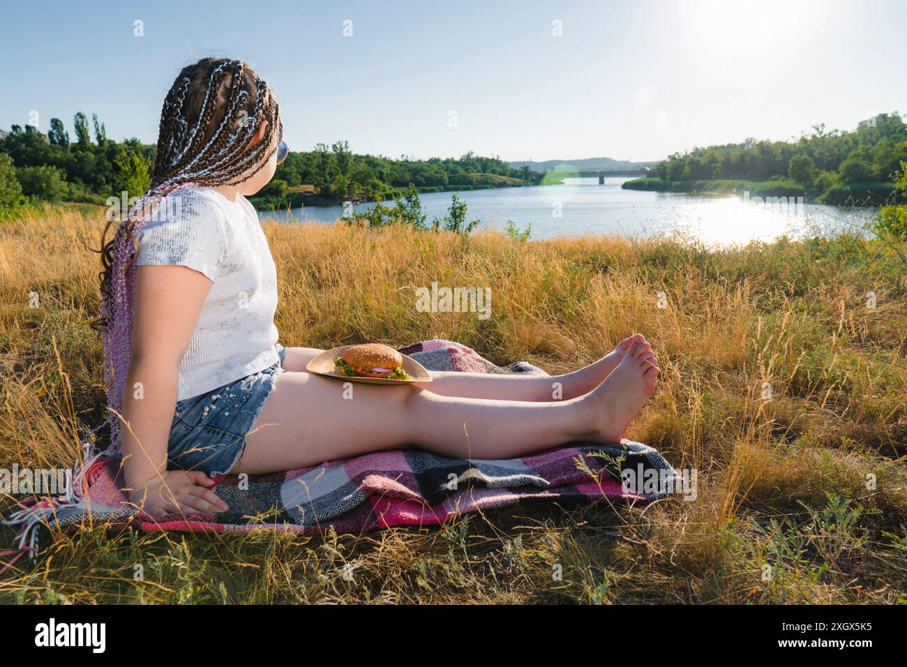 Ein Mädchen mit einem Burger sitzt an einem sonnigen Sommertag am Flussufer. Auf Dem Land. Picknick im Freien Stockfoto