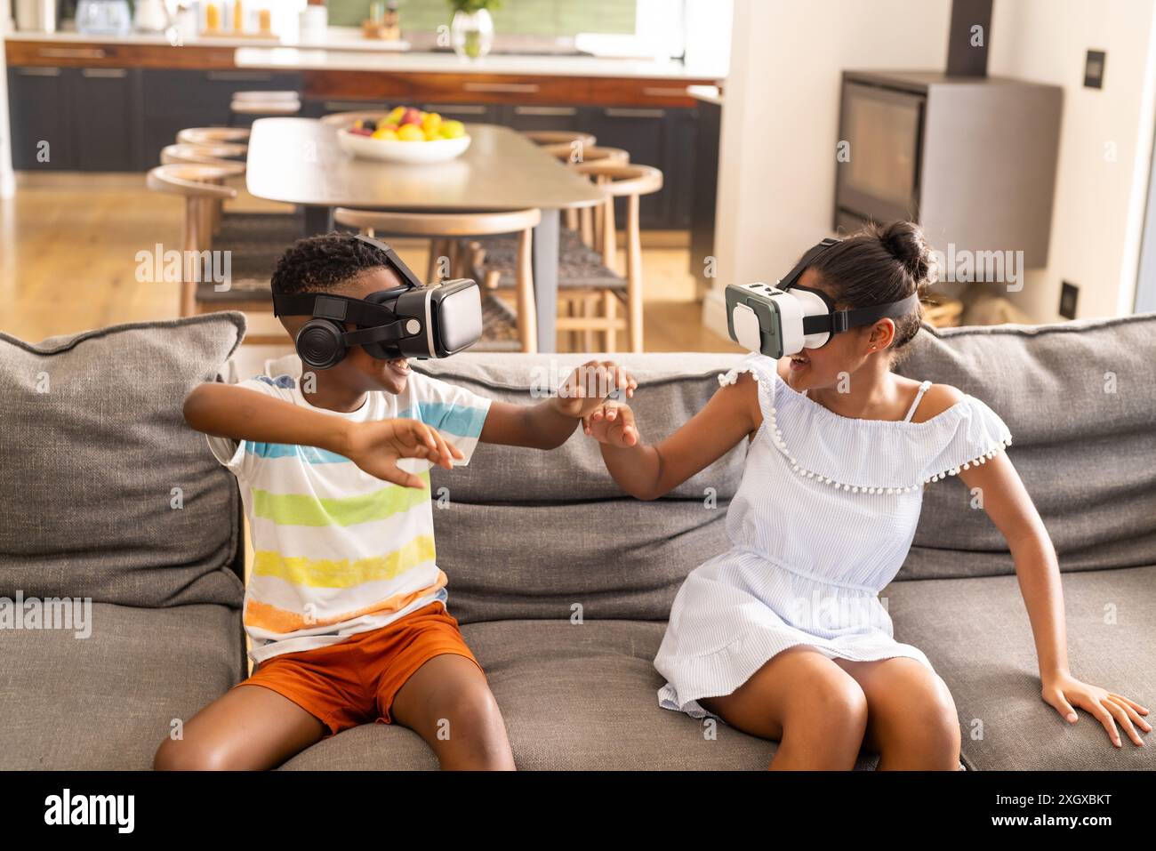Birassische Geschwister genießen VR-Spiele zu Hause. Sie haben Spaß mit Virtual-Reality-Technologie in einem gemütlichen Wohnzimmer. Stockfoto