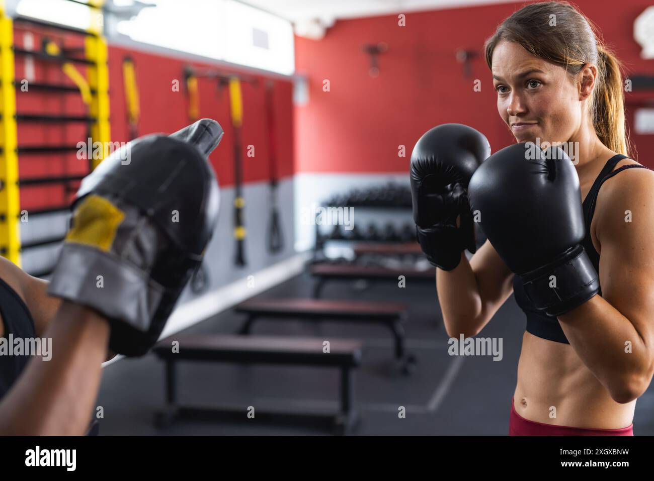 Eine fitte junge Kaukasierin trainiert im Fitnessstudio im Boxen. Sie ist konzentriert, während sie mit einem Afroamerikaner trainiert, um ihre Fähigkeiten zu verbessern. Stockfoto