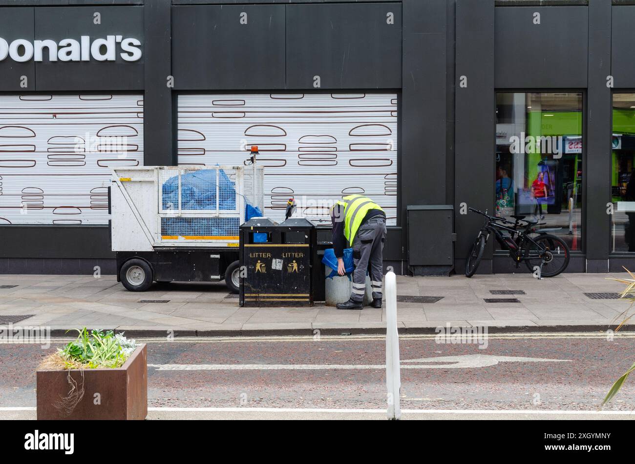 Belfast, County Antrim Nordirland 05. Juli 2024 - Gemeindeangestellter leert Abfalleimer auf einer Straße im Stadtzentrum vor einem großen Einzelhandelsgeschäft Stockfoto