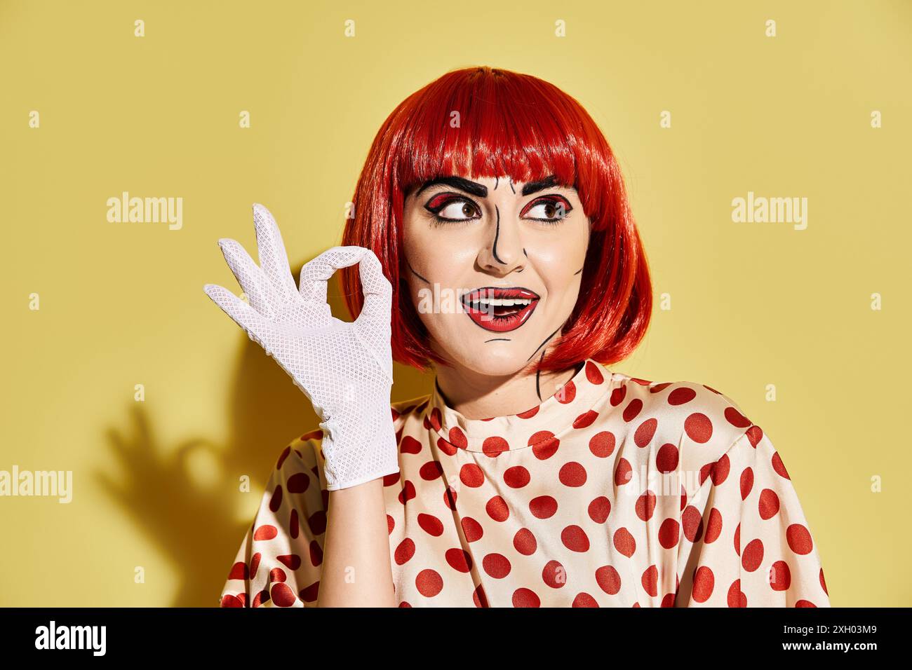 Eine auffällige rothaarige Frau mit Pop-Art-Make-up trägt ein Polka-Dot-Shirt und weiße Handschuhe auf gelbem Hintergrund. Stockfoto