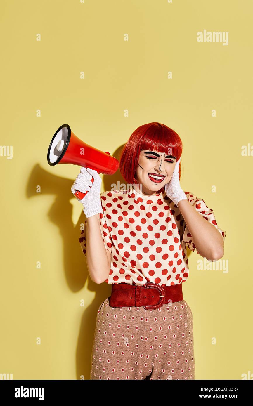 Eine leuchtende rothaarige Frau in einer Punktbluse hält selbstbewusst ein rot-weißes Megaphon auf gelbem Hintergrund. Stockfoto