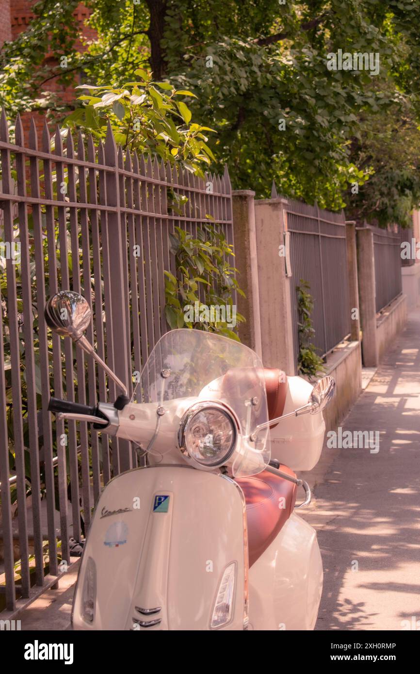 Eine weiße Vespa, die in einer sonnendurchfluteten Straße neben einem Zaun und Bäumen parkt, Mailand, Italien Stockfoto