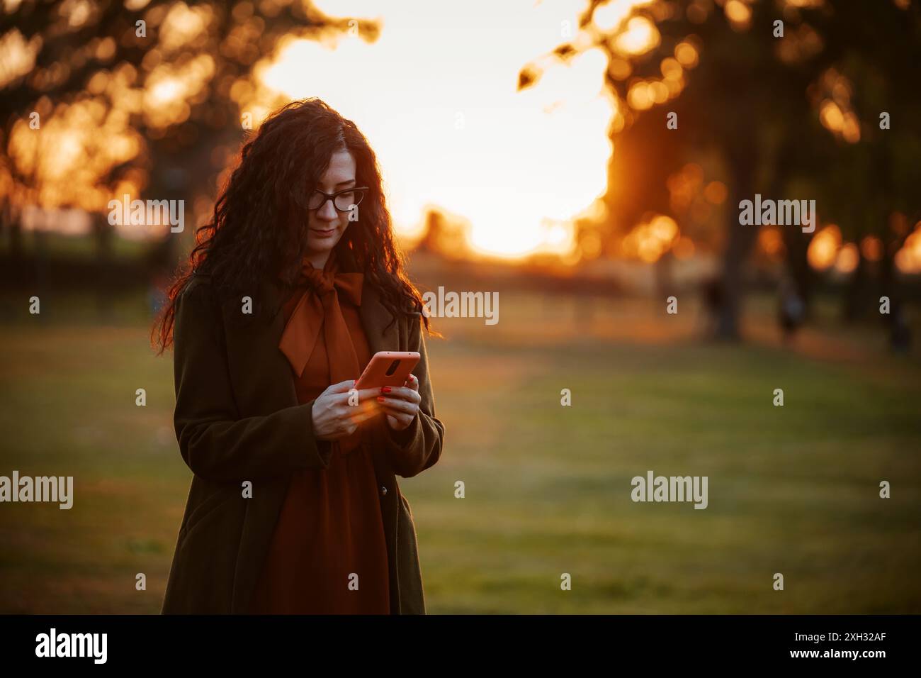 Eine Frau mit langen lockigen Haaren und einer Brille überprüft ihr Telefon, während sie im Park steht und im warmen Licht der untergehenden Sonne gebadet ist. Stockfoto