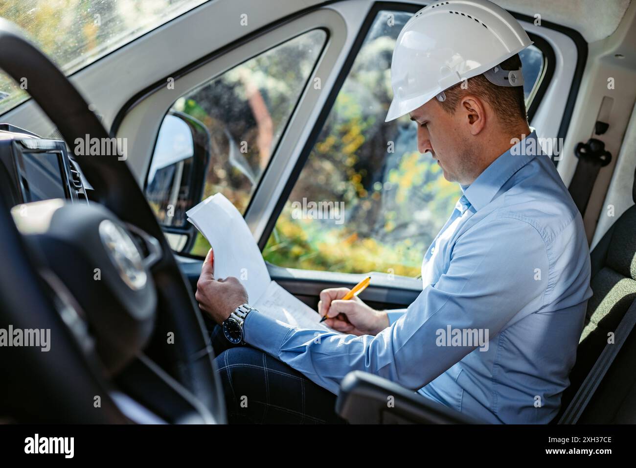 Ein Bauarbeiter mit Schutzhelm sitzt in einem weißen Van und prüft Dokumente und schreibt Notizen. Stockfoto