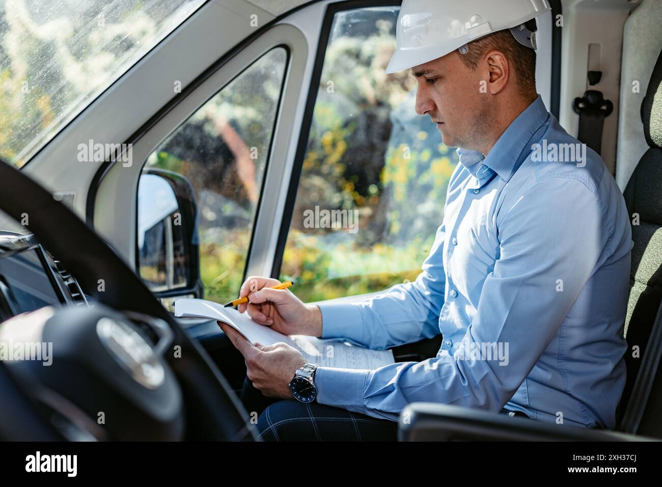 Ein Bauarbeiter mit weißem Schutzhelm sitzt auf dem Fahrersitz eines Lieferwagens und schreibt mit einem gelben Bleistift in ein Notizbuch. Stockfoto
