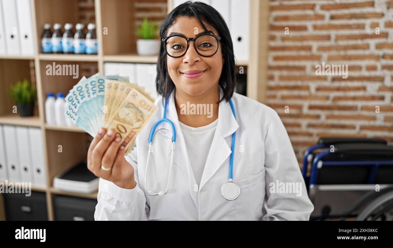 Eine lächelnde Ärztin in einem weißen Mantel, die brasilianische Währung in einer Klinik hält, die Gesundheitskosten oder Gehalt darstellt. Stockfoto