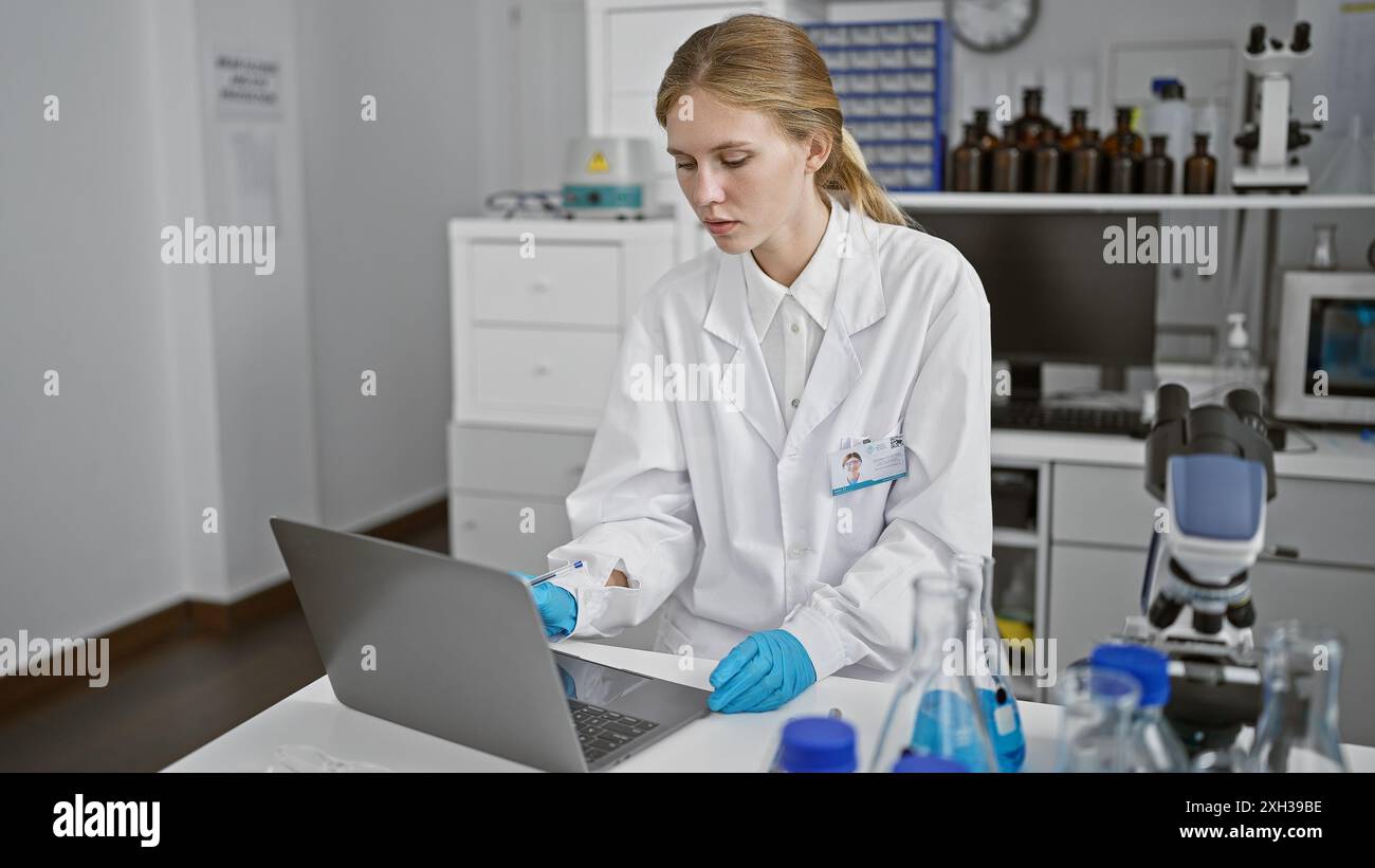 Eine konzentrierte blonde Frau in einem weißen Labormantel arbeitet an einem Laptop in einer modernen Laborumgebung. Stockfoto