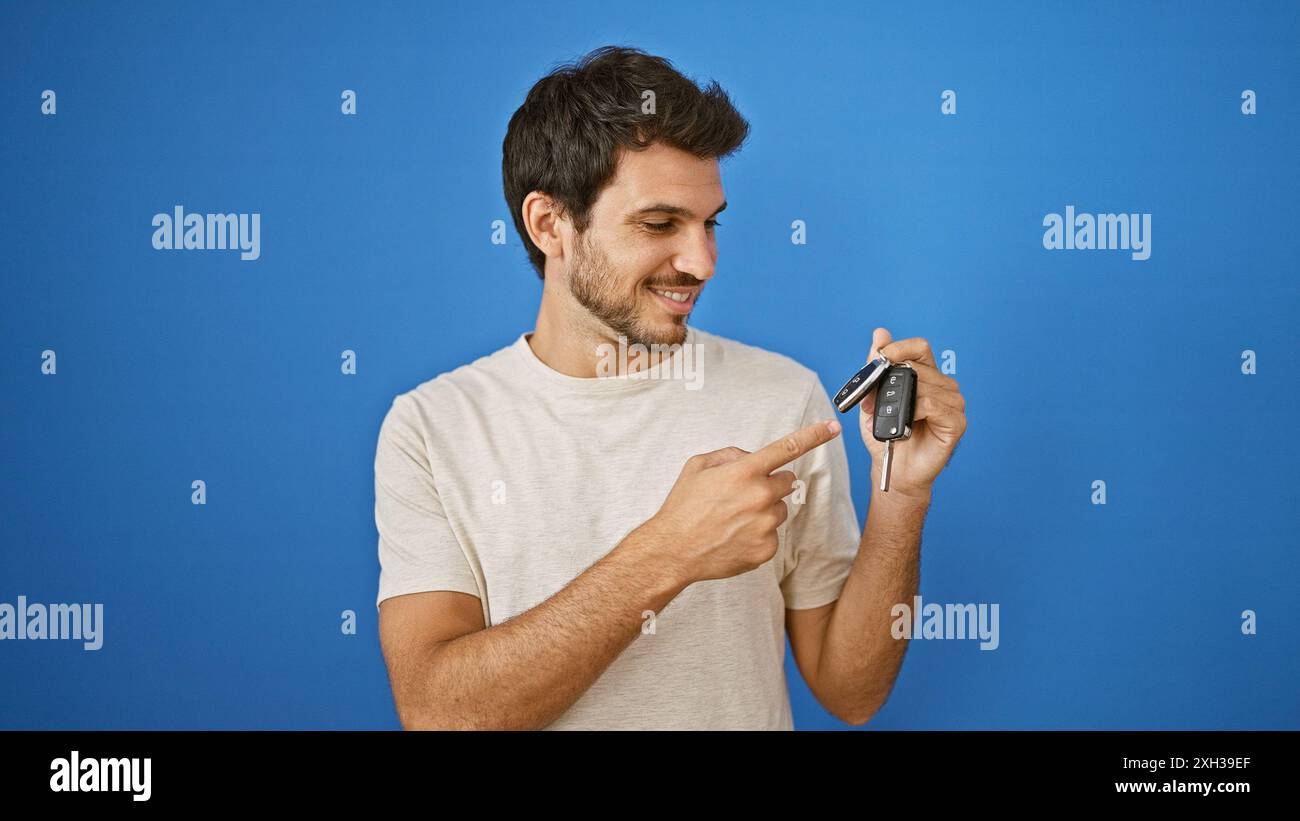 Hübscher junger hispanischer Mann, der lächelnd auf eine Schlüsselfernbedienung des Autos zeigt, an einer blauen Wand draußen. Stockfoto