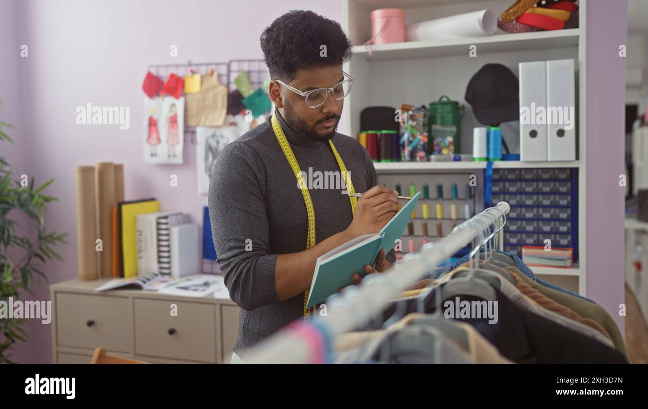afroamerikanischer Schneider macht Notizen in einem farbenfrohen Atelier, umgeben von Stoffen und Designskizzen. Stockfoto