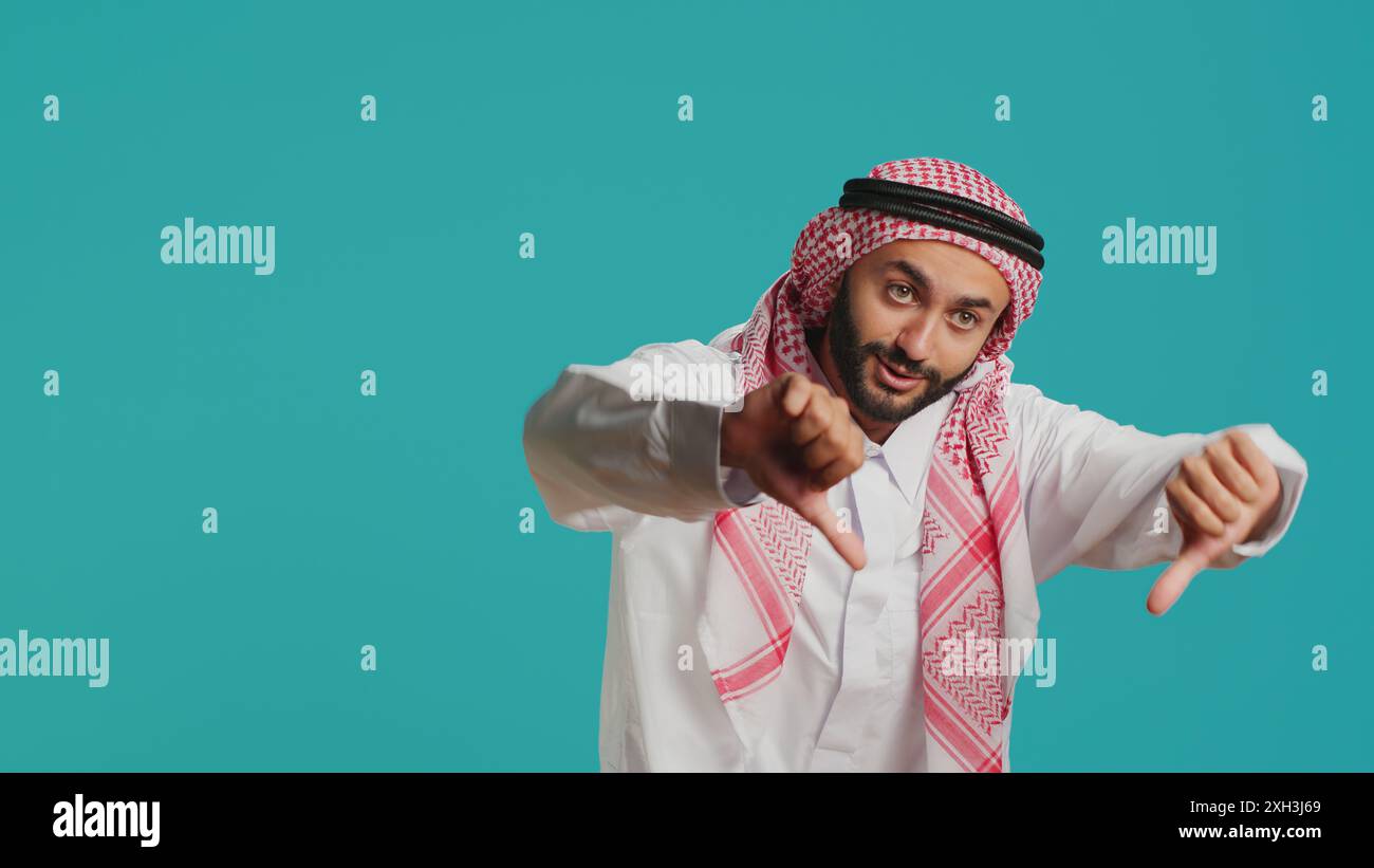Eine arabische Person in historischem Kleid und Kufiyah gibt die Daumen nach unten und zeigt Unzufriedenheit mit dem, was sie sieht. Im Studio zeigt islamische Erwachsene eine deutliche Ablehnung und ein negatives Signal. Stockfoto