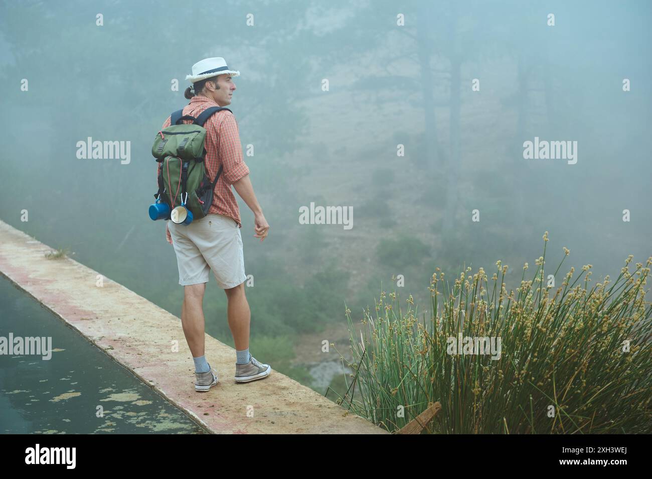 Mann mit Hut und orangefarbenem Hemd, mit Rucksack, stehend auf einem Weg in einer nebeligen Waldumgebung. Konzept: Abenteuer, Natur, Erkundung. Stockfoto