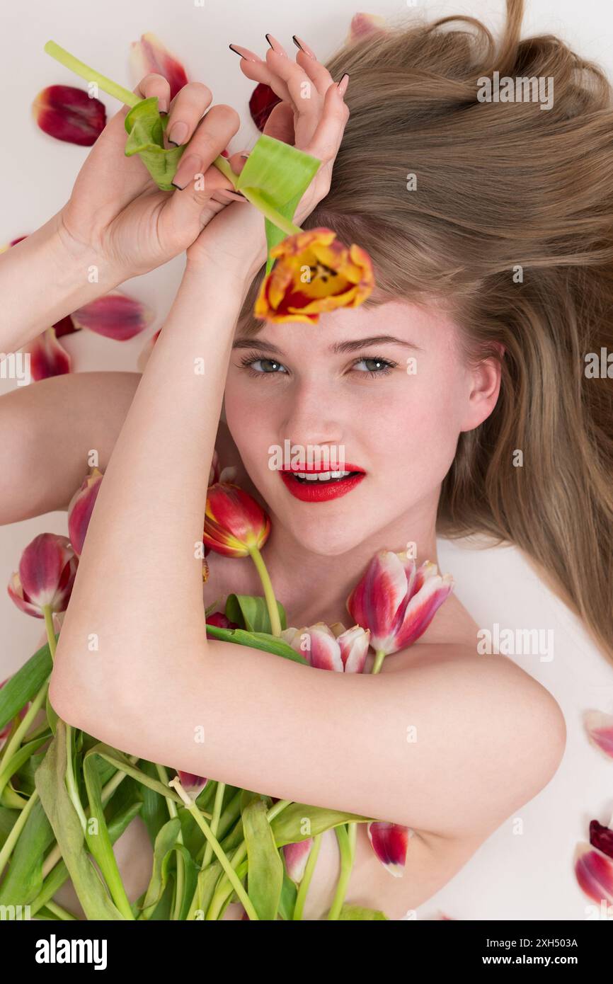 Schöne blonde Dame, die mit einem Strauß Frühlingstulpen zum Valentinstag liegt und ihre Brust bedeckt. Die wunderschöne junge Blondine schaut liebevoll in die Kamera Stockfoto