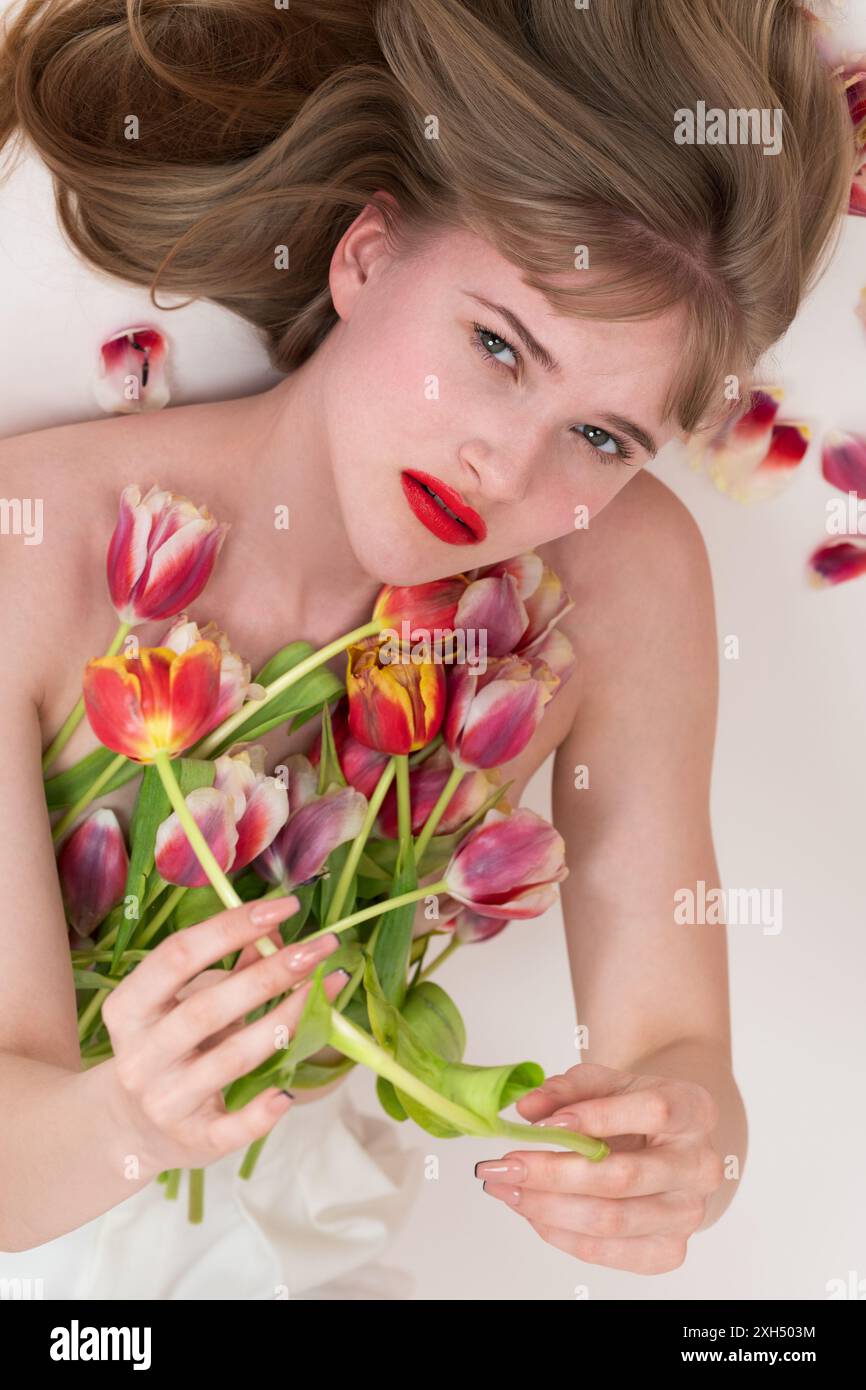 Junge Frau mit Frühlingstulpen, die auf der Brust liegen und sie bedecken, in die Kamera schauen. Blonde Frau, die Blumen hält, während sie sich auf weißem Hintergru legt Stockfoto
