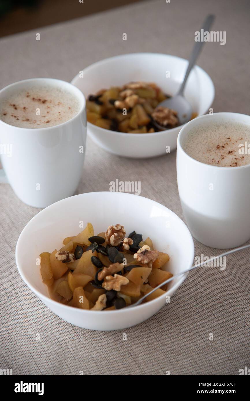 Ein gemütliches Frühstück mit zwei weißen Tassen schaumigen Cappuccino und zwei Schüsseln gebackenen Apfeln, Nüssen und Kürbiskernen auf einer beigen Tischdecke Stockfoto