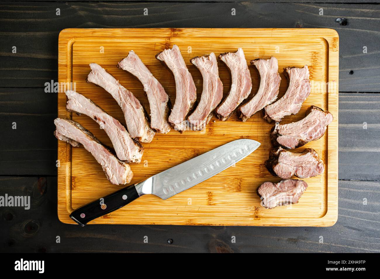 Gekochte Schweinerippchen, die auf einem Schneidebrett aufgefächert werden: Geröstete Baby-Back-Rippchen ohne Sauce, die auf einem Bambusschnitzbrett mit einem Kochmesser geschnitten wurden Stockfoto