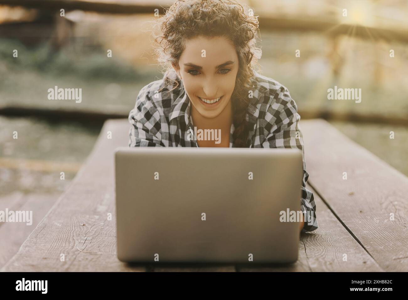 Eine junge Frau mit lockigen Haaren in einem Zopf, gekleidet in einem karierten Hemd, liegt mit einem Laptop auf ihrem Bauch. Sie ist tief in ihre Arbeit vertieft, mit Th Stockfoto