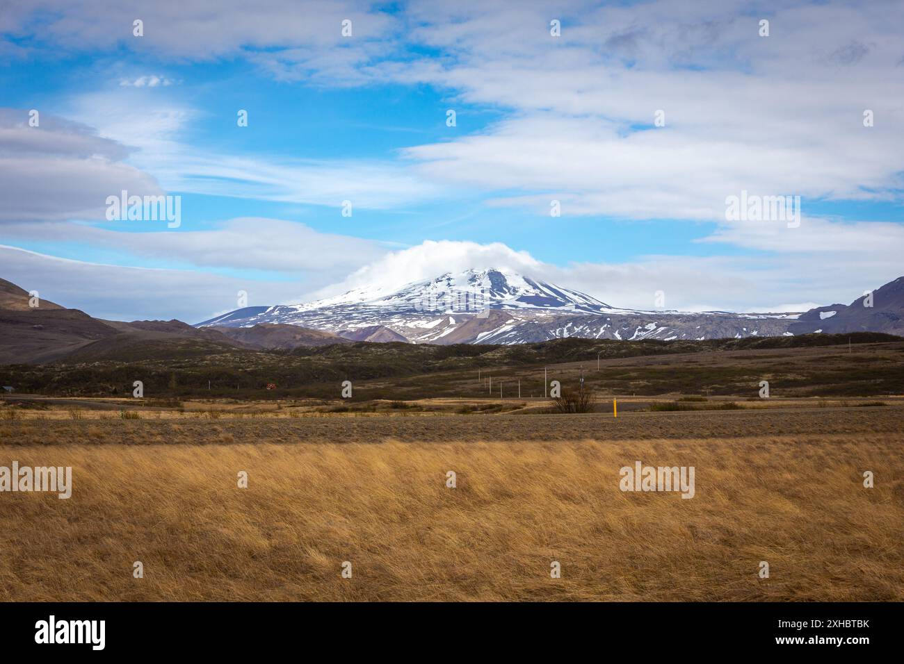 Isländische Landschaft mit Grasland, Lavafeldern und schneebedecktem Hekla-Vulkan mit Wolken und blauem Himmel. Stockfoto