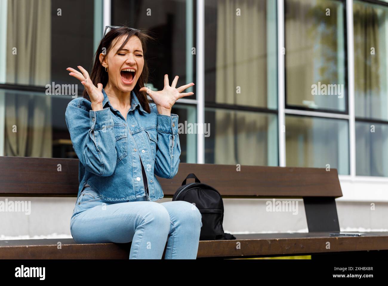 Eine junge Frau in einer Jeansjacke sitzt auf einer Bank und schrie überrascht mit erhobenen Händen. Stockfoto