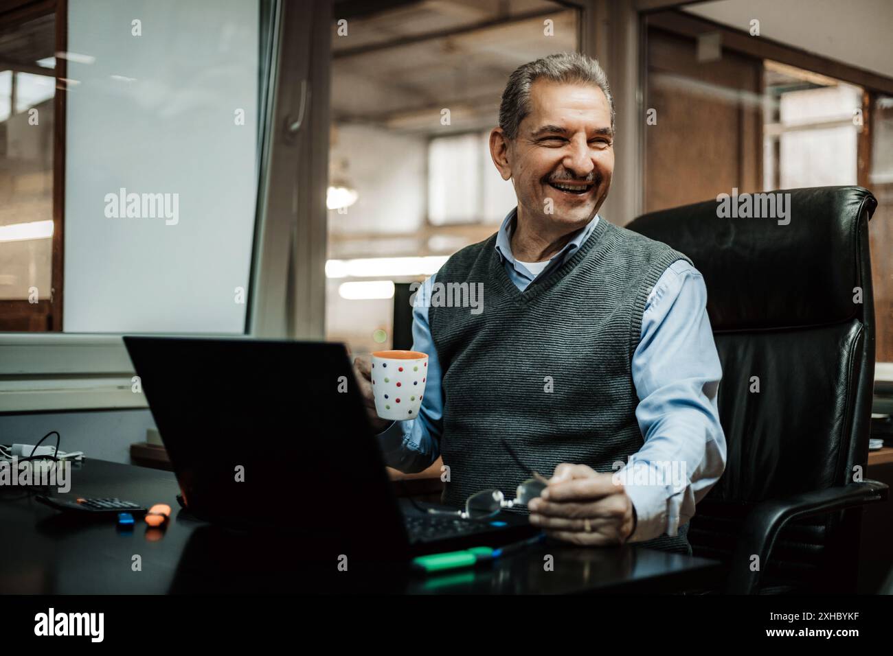 Ein Mann sitzt an seinem Schreibtisch in einem Büro und lächelt, während er nach rechts blickt. Er hält einen Becher in der Hand und schaut sich etwas aus der Kamera an. Stockfoto
