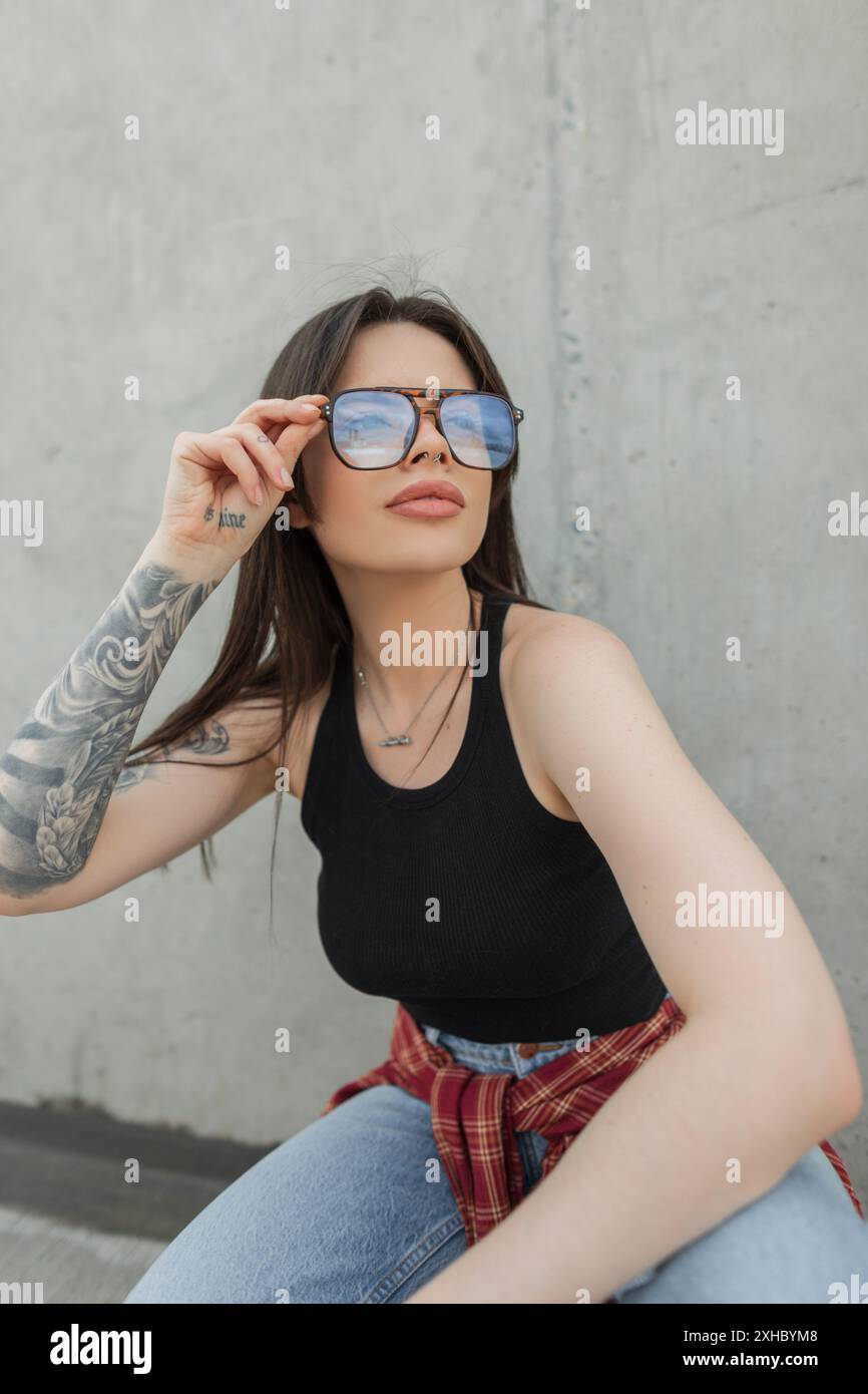 Cooles, schönes, modisches Studentenmädchen mit einer Brille mit Tattoos in modischer Kleidung, die auf der Straße posiert Stockfoto