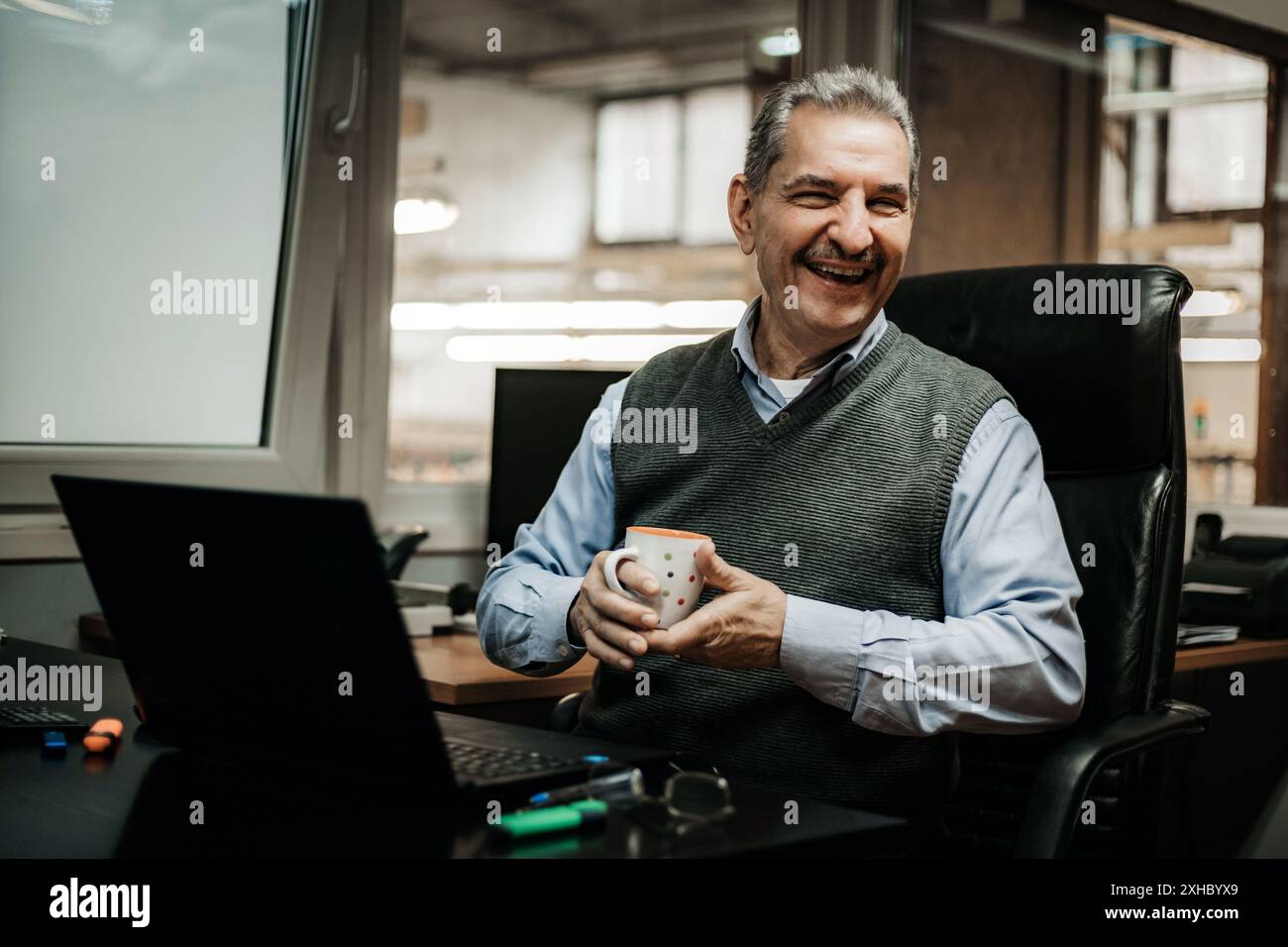 Ein lächelnder Seniorenmann sitzt an seinem Schreibtisch in einem Büro und hält einen Becher in der rechten Hand. Er schaut in die Kamera und scheint seine Arbeit zu genießen. Stockfoto